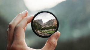 Imagem de uma pessoa segurando uma lente de aumento e uma paisagem natural vista atraves dela